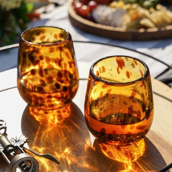 Two amber solid cups with a spotted plan are placed connected a woody table, reflecting light. A vino opener is nearby, with snacks blurred successful the background