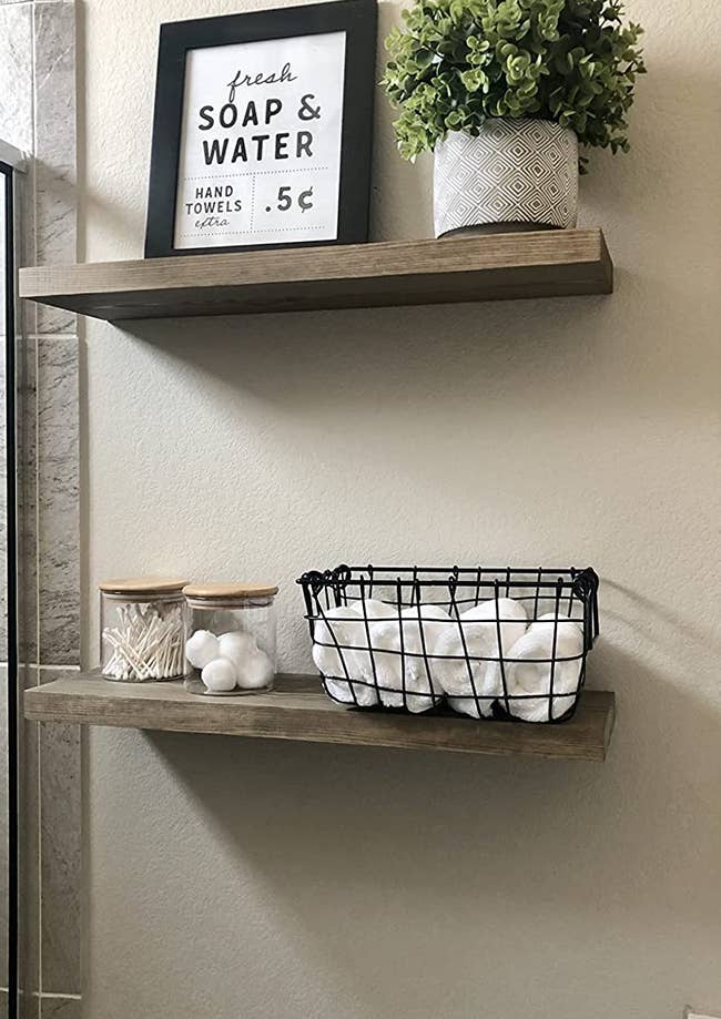 Reviewer image of two wall-mounted wooden shelfs with basket and containers of toiletries and towels 