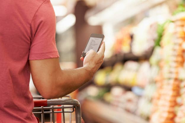A person is seen from behind, holding a phone with a shopping list while pushing a shopping cart in a grocery store aisle