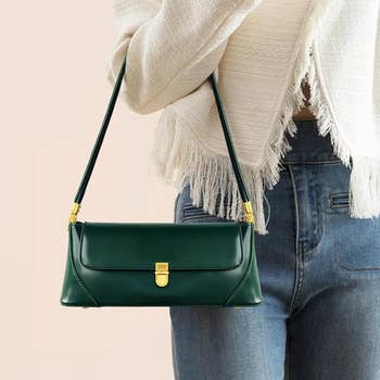 Person holding a green handbag with a structured design and gold clasp, paired with a fringed jacket and jeans
