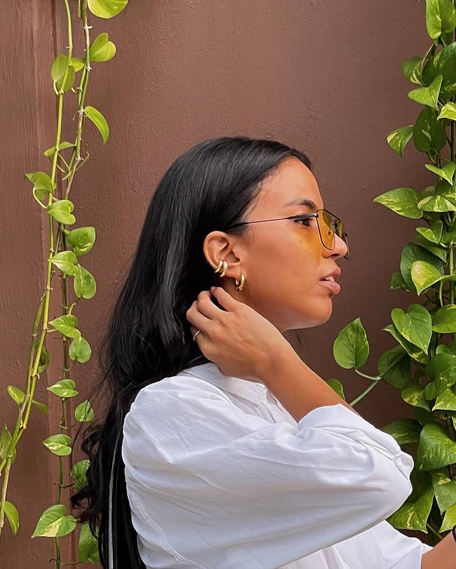Person with sunglasses and hoop earrings, wearing a achromatic blouse, stands against a leafy background. Ideal for manner oregon accessory buying context
