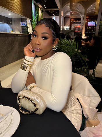 Person in an elegant long-sleeve dress, adorned with statement jewelry, sits at a dining table with a stylish clutch bag