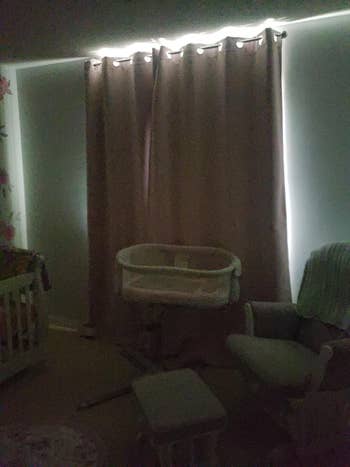 Dimly lit nursery with blackout curtains. A cradle, rocking chair, and footstool are visible, suggesting a cozy, sleep-friendly setup