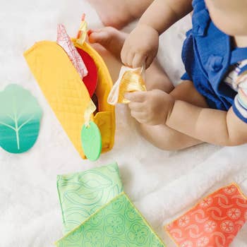 Baby playing with a soft, taco-shaped artifact  with smiling faces, cloth  fillings, and colorful felt pieces, connected  a brushed  surface