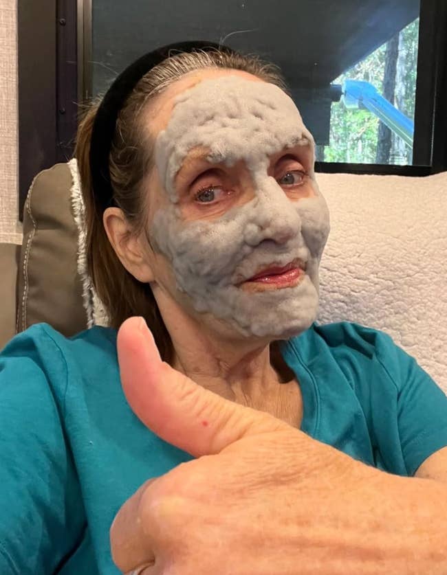 Person with a face mask giving a thumbs-up, wearing a teal shirt and black headband, seated on a couch; beauty or skincare routine theme