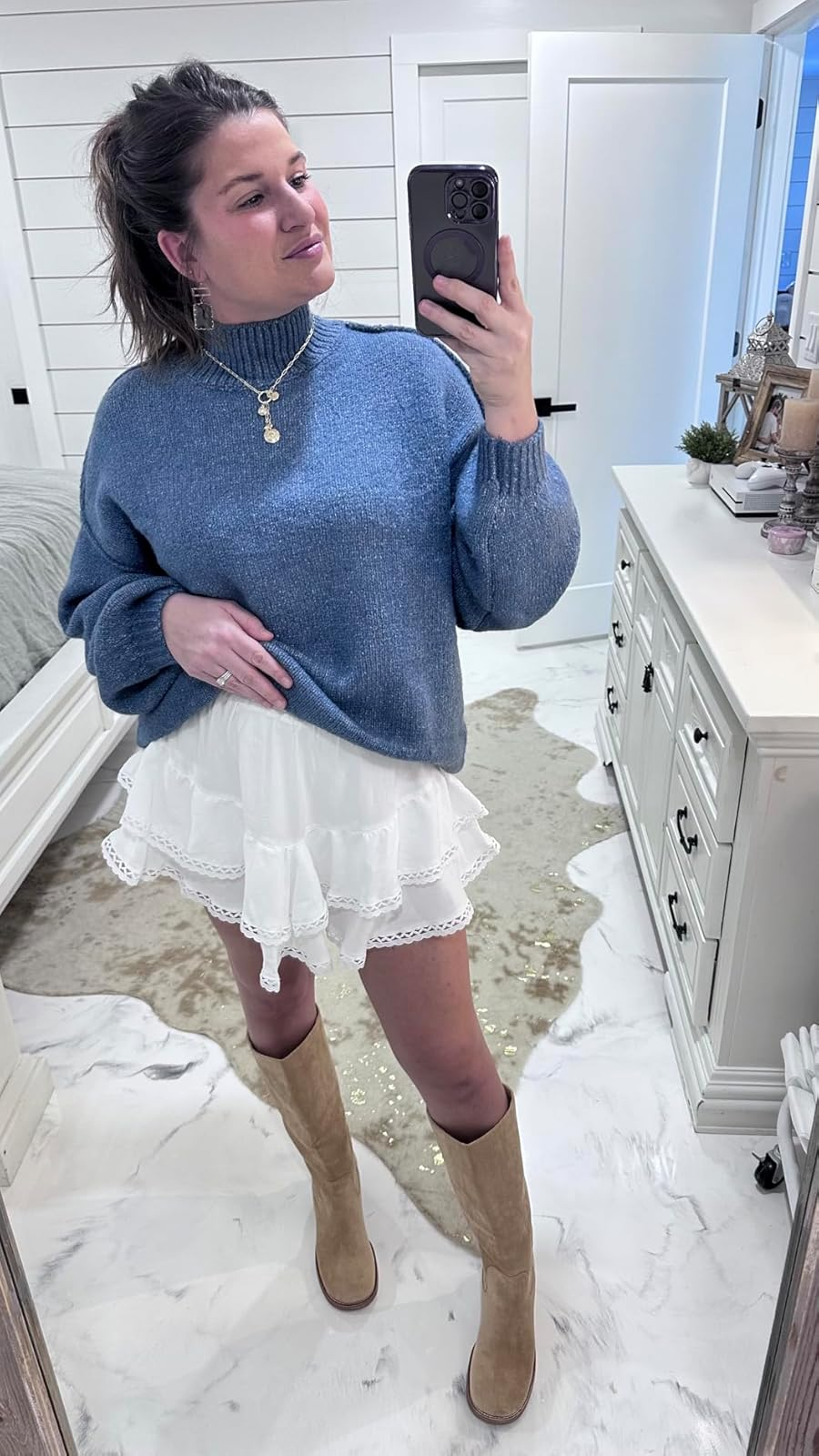 Person in a cozy sweater, skirt, and knee-high boots takes a mirror selfie in a stylish bedroom