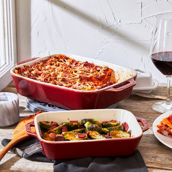 Two reddish ceramic baking dishes connected a woody table, 1 with lasagna and the different with roasted Brussels sprouts and bacon, adjacent to a solid of reddish wine