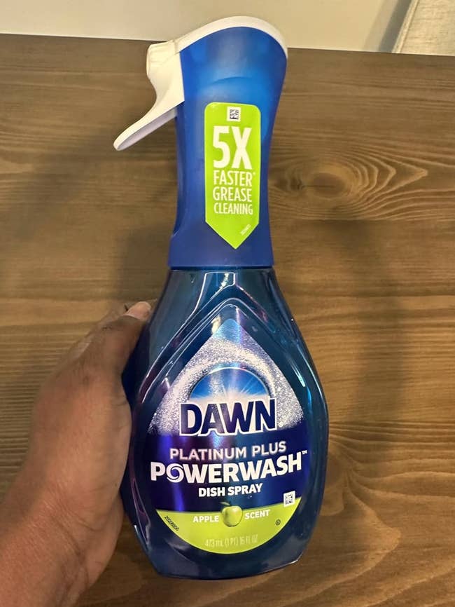 A manus  holds a vessel  of Dawn Platinum Plus Powerwash Dish Spray with Apple scent, known for 5x faster grease cleaning, connected  a woody  table