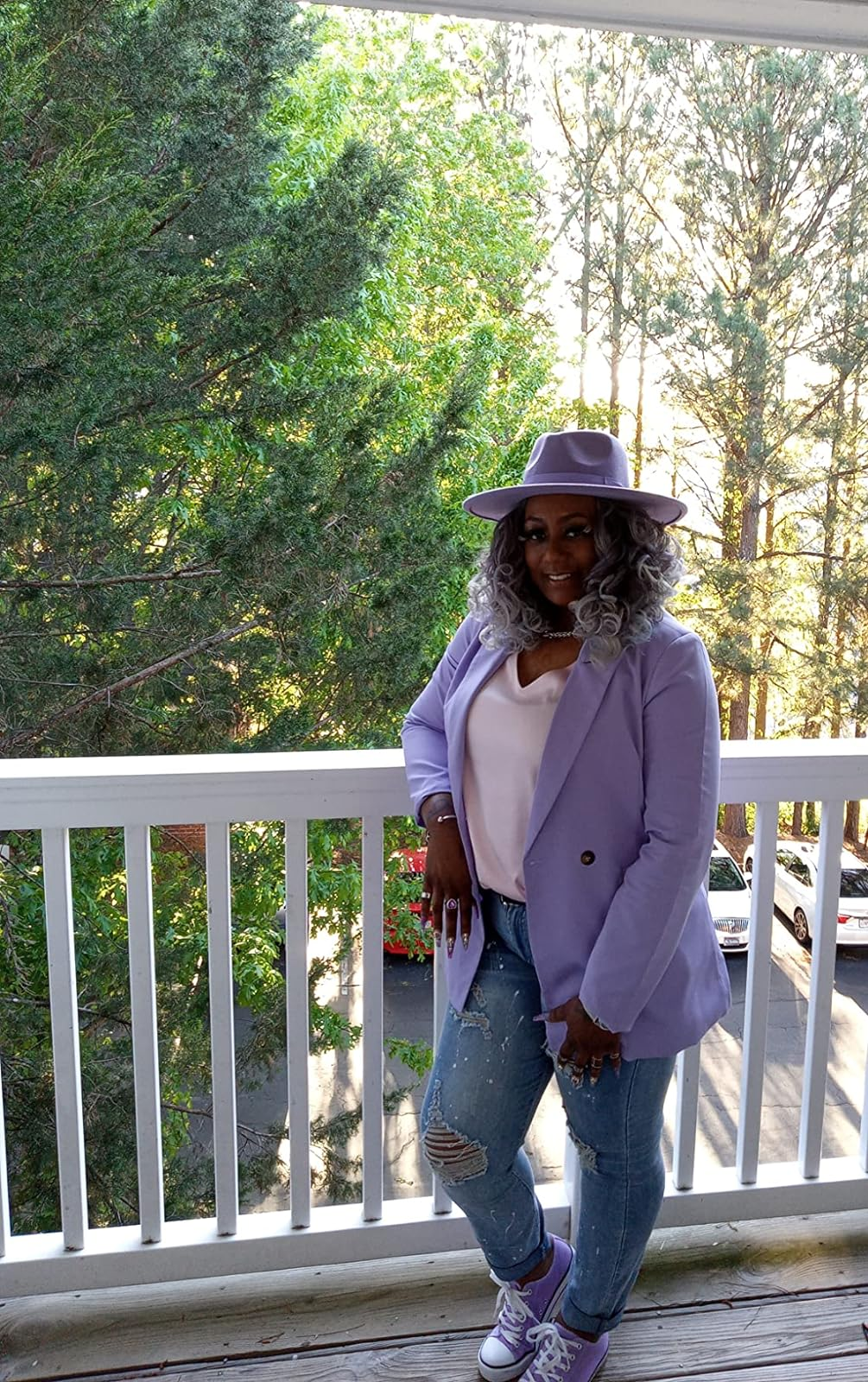 Person standing on a balcony, wearing a stylish lavender blazer, matching hat, white top, distressed jeans, and sneakers with trees in the background