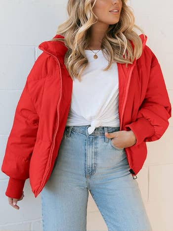 Person wearing a casual red puffer jacket over a white knotted t-shirt and light blue jeans, posing against a white wall