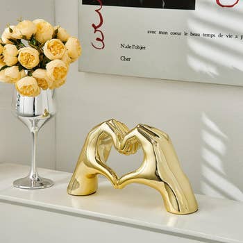 Gold-toned sculpture of two hands forming a heart, on a white shelf next to a vase of yellow roses