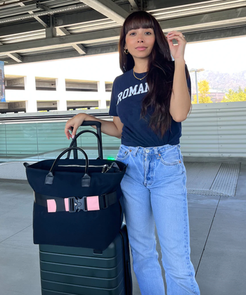 model standing next to a green suitcase with a black carry-on strapped to the handle using a pink and black belt