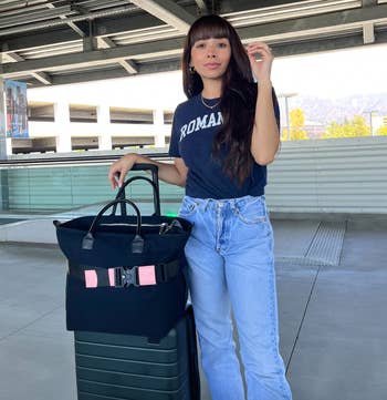 model standing next to a green suitcase with a black carry-on strapped to the handle using a pink and black belt