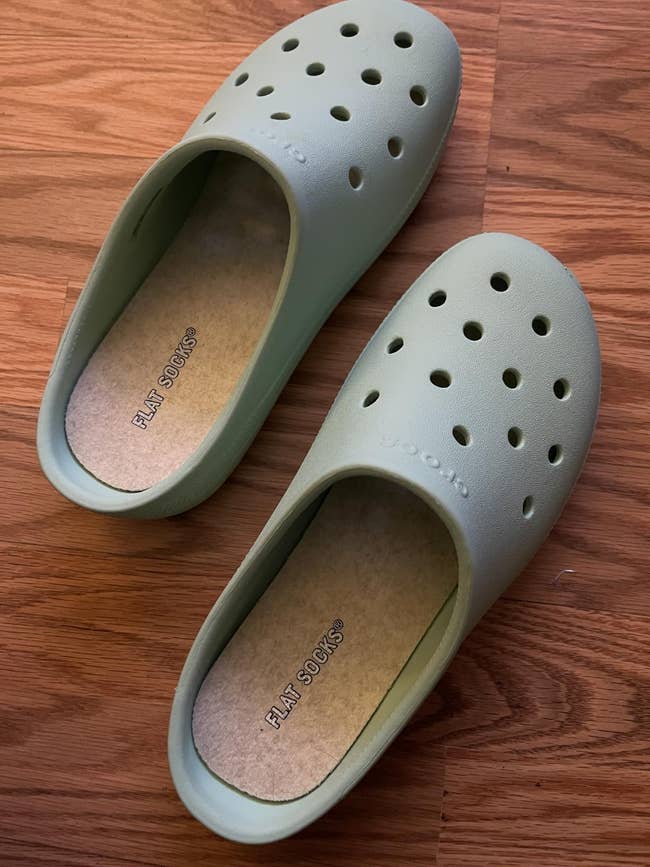Two slip-on shoes with perforated tops placed connected a wood surface, featuring branded insoles labeled