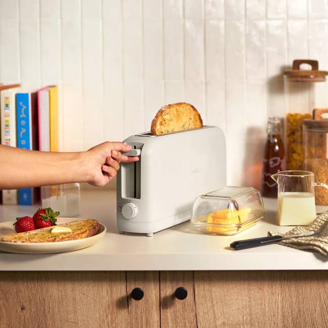 A person uses a sleek toaster with toast popping out, on a kitchen counter set with butter, strawberries, and milk