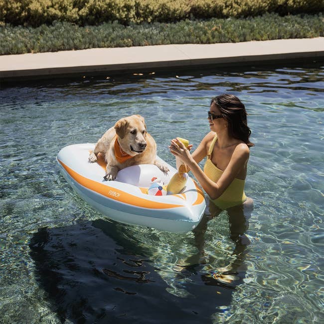 white and orange pool float sized for a dog with a yellow lab chilling on it in water with a model next to it