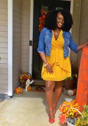 Woman in yellow dress and denim jacket posing by a door with fall decorations