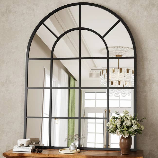 Arched grid mirror on a wooden table with books, a small vase, and flowers, reflecting a chandelier and French doors in the background