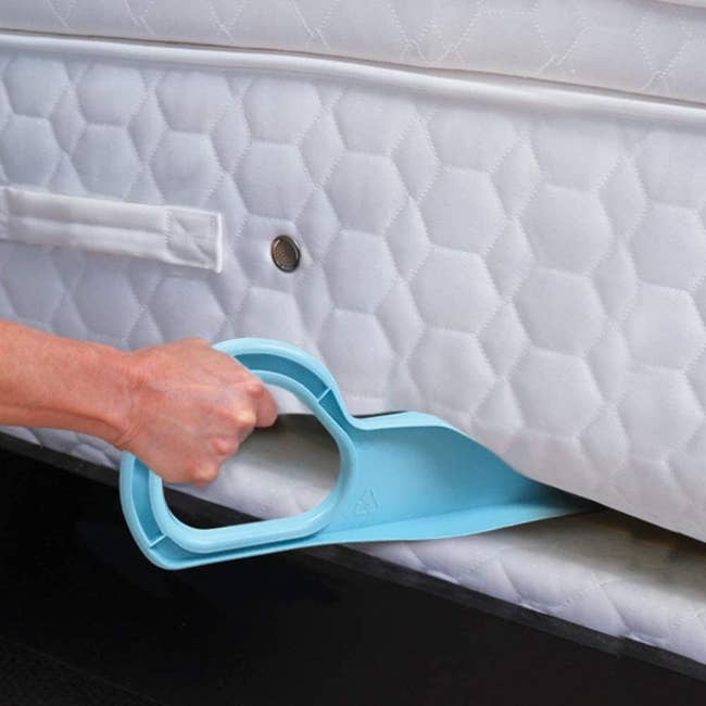 A hand using a blue tool to lift a mattress, demonstrating an easy bed-making accessory