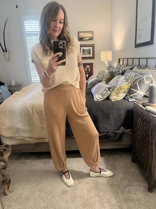Person successful a cozy bedroom, smiling, wears a sleeveless top, relaxed pants, and slip-on shoes, with a canine astatine their feet