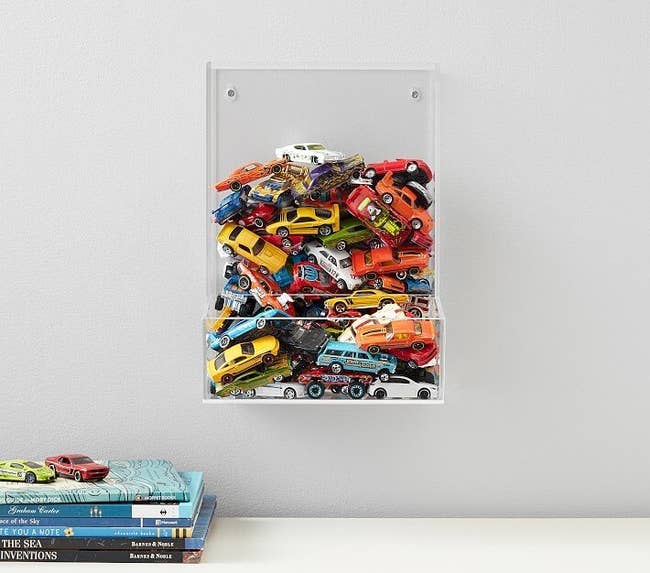 clear cubby shelf mounted to wall with opening to pull toys from. filled with tiny toy cars. 