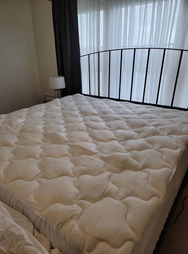 Plush, quilted mattress topper on a bed in a sunlit room with sheer curtains and a metal headboard