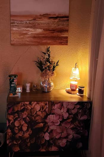 Meja rias kecil dengan desain bunga, dihiasi dengan vas bunga, lampu, lilin, dan berbagai barang kecil, di bawah lukisan lanskap