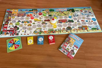 Children&rsquo;s board game set up on a wooden floor, featuring a colorful game board, spinner, cards, and instruction manual. The game theme is busy-town related