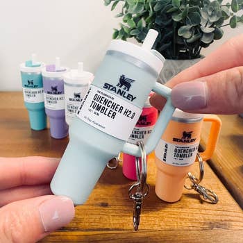 Hand holding a keychain modeled after the Stanley Quencher H2.0 tumbler, with full-sized tumblers in the background