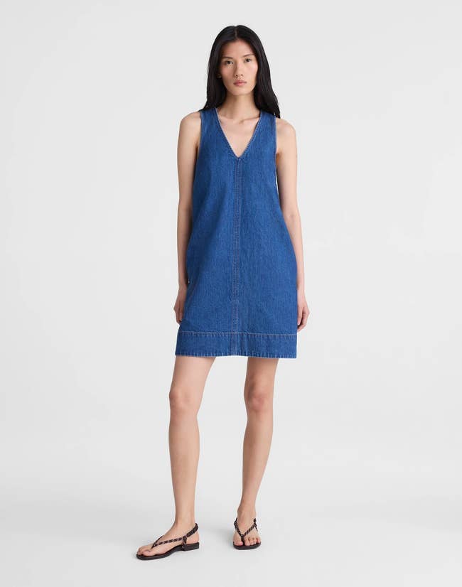 Person in a sleeveless denim dress and sandals poses against a plain background