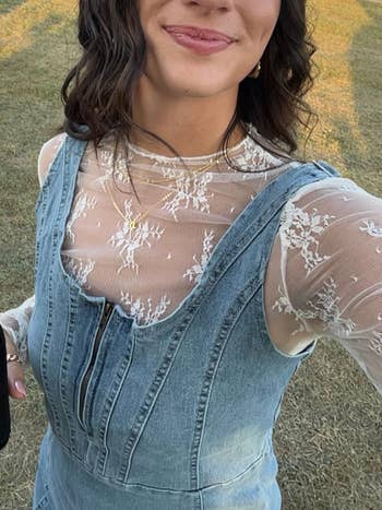 Person wearing a lace top under a denim dress with a front zipper, holding a black object in one hand, standing on grass