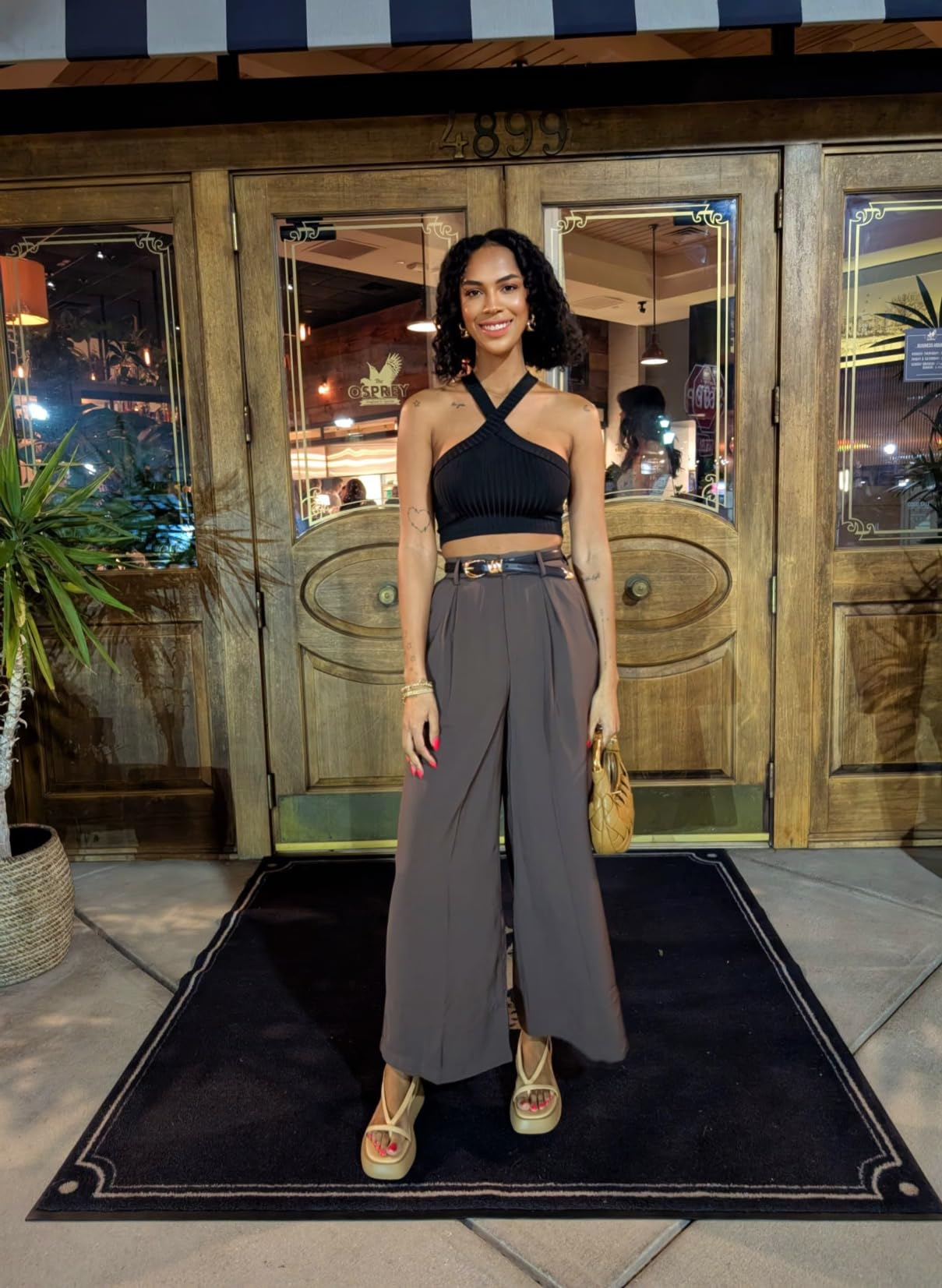 Person successful  chic, casual outfit with a achromatic  halter apical  and wide-leg pants lasting  extracurricular  a rustic store  entrance, holding a tan container  and wearing sandals