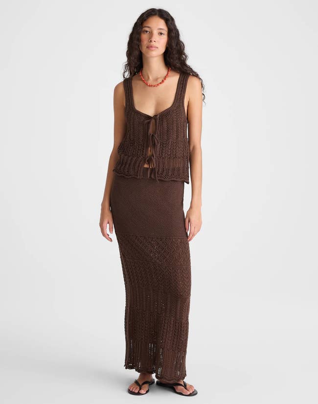 Model wearing a sleeveless, open-knit top and long skirt with a crochet pattern, paired with sandals and a red necklace