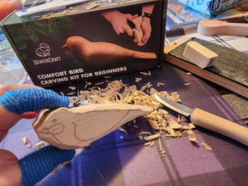 Bird carving kit with a wooden block, carving shavings, and a carving tool on a table. A hand in a protective glove holds a carved bird shape
