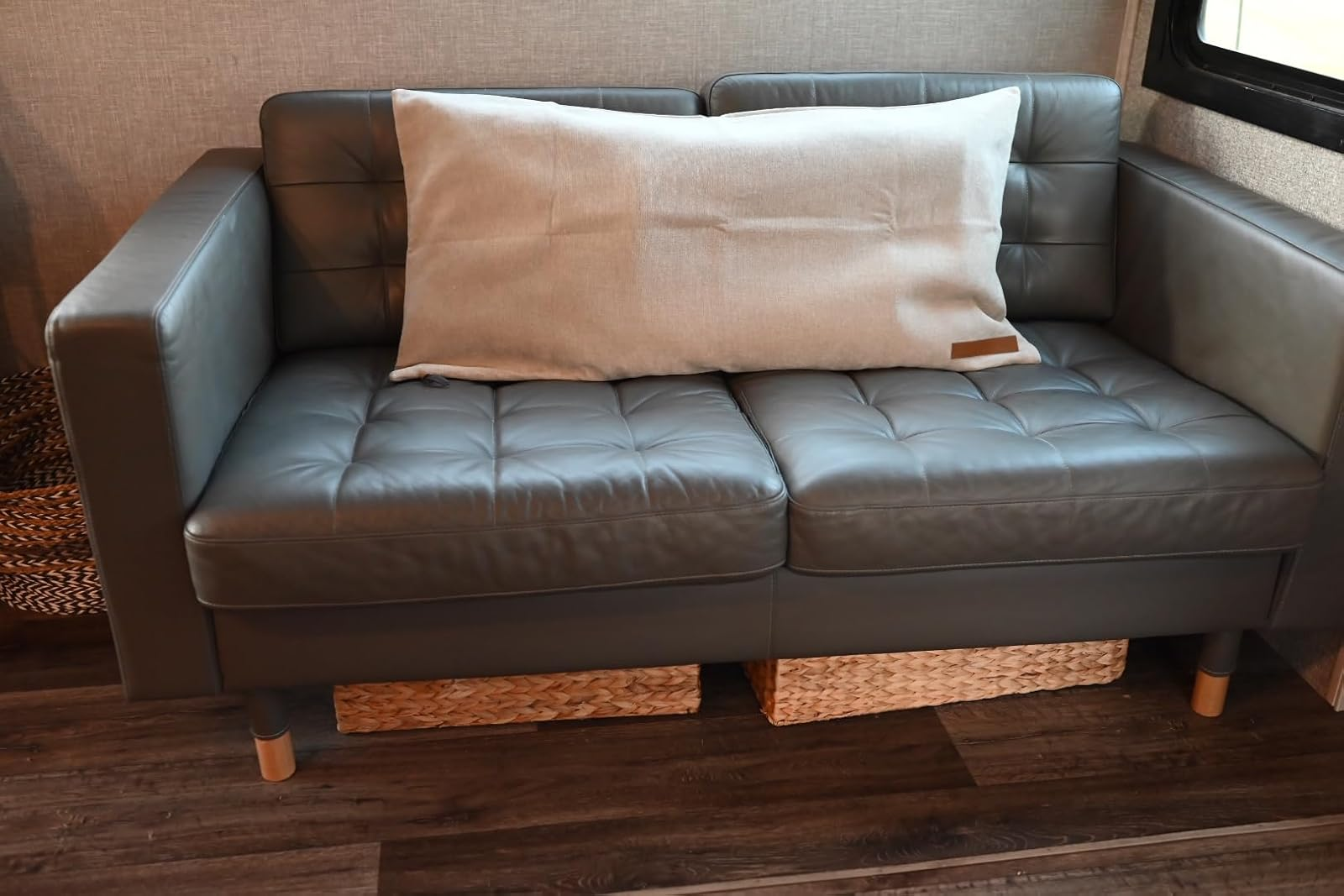 Modern gray leather loveseat with a large cushion and woven baskets underneath, set on wooden flooring. Ideal for space-saving living areas