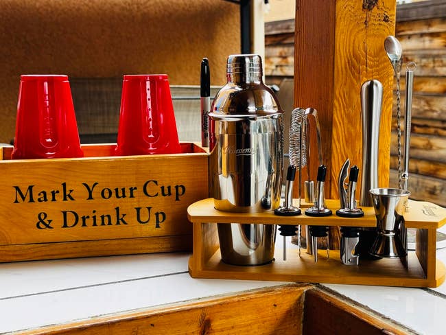 Bar acceptable with shaker, jigger, and tools successful a woody holder beside a container with reddish cups labeled
