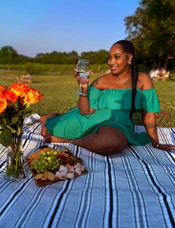 reviewer in off-shoulder dress enjoying a picnic with a glass of wine, fruits, and cheese on a blanket outdoors
