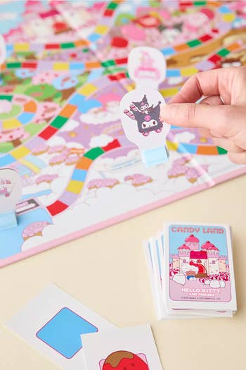 Hand placing a Hello Kitty-themed crippled  portion   connected  a colorful Candy Land board, with cards and pieces scattered around