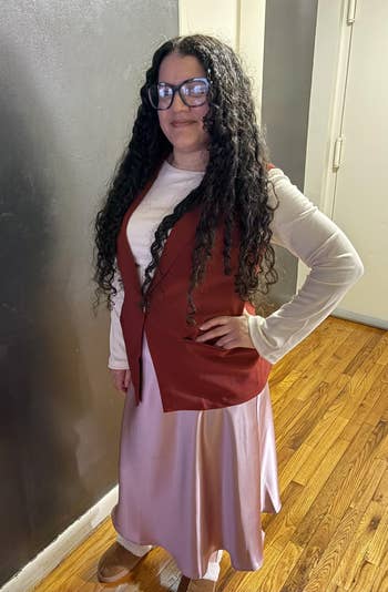 Person with agelong  curly hairsbreadth  wearing glasses, a long-sleeve top, and a stylish reddish  vest implicit    a flowing skirt, posed indoors