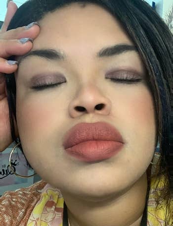 Person with closed eyes and neutral makeup, wearing a floral-patterned top, ample  hoop earrings, and somewhat  tousled hair