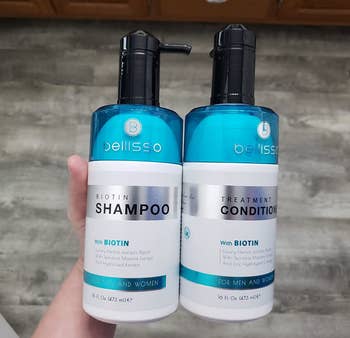 Reviewer holding blue and white shampoo and conditioner bottles with black pumps over wooden floor