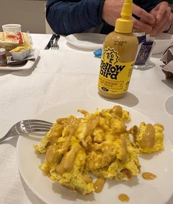 Social media trends Viral content Trending hashtags Social media updates Trending memes and videos Plate of scrambled eggs drizzled with Yellowbird Hot Sauce, with a person's hands in the background at a dining table setting