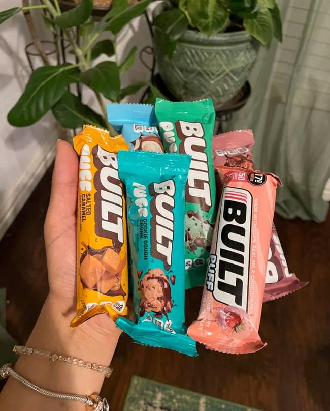 A hand holds various Built protein bars, including flavors like Salted Caramel, Cookie Dough, and Strawberry, displayed in a casual home setting
