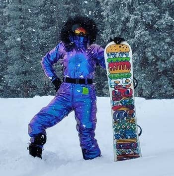 reviewer wearing the black belt around the waist of their blue snowsuit while standing next to a snowboard in the snow