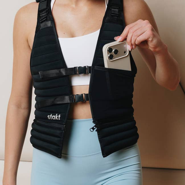 Person wearing a weighted vest, placing a smartphone in a front pocket, and wearing a sports bra and leggings