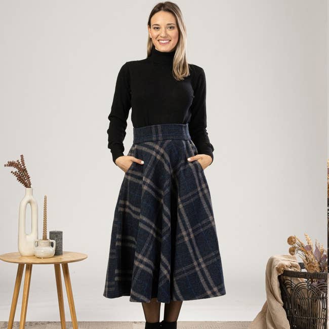 Woman wearing a black turtleneck and a high-waisted, plaid, full midi skirt with pockets, standing indoors next to a small table and decorative items
