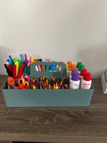 A teal organizer holding various school supplies including markers, crayons, scissors, and paint bottles
