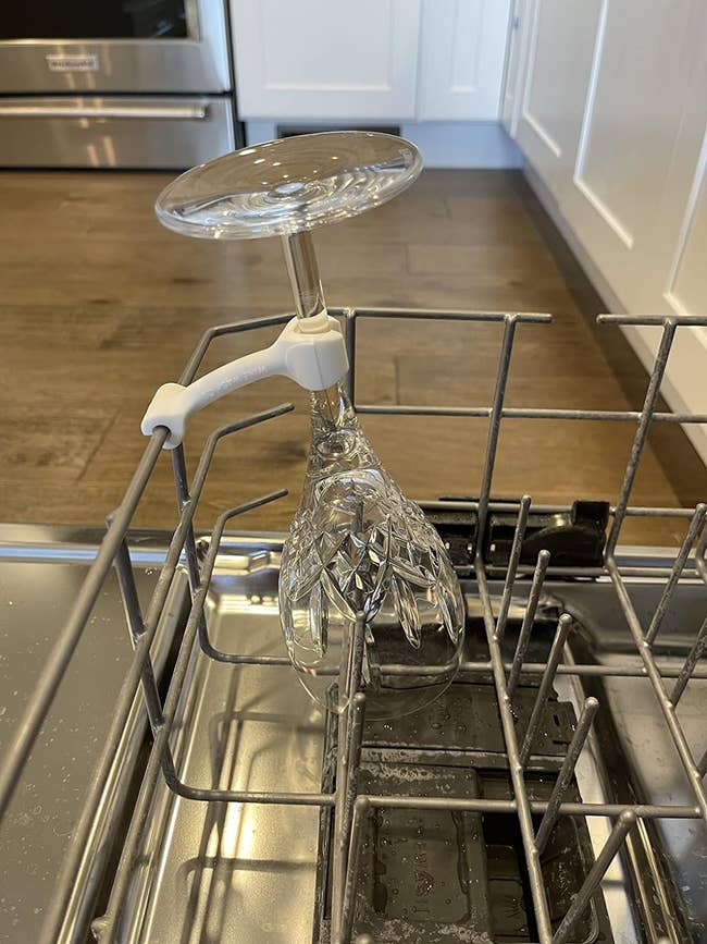 A clear wine glass is securely placed upside down in a dishwasher rack using a white plastic glass holder attached to a metal rod