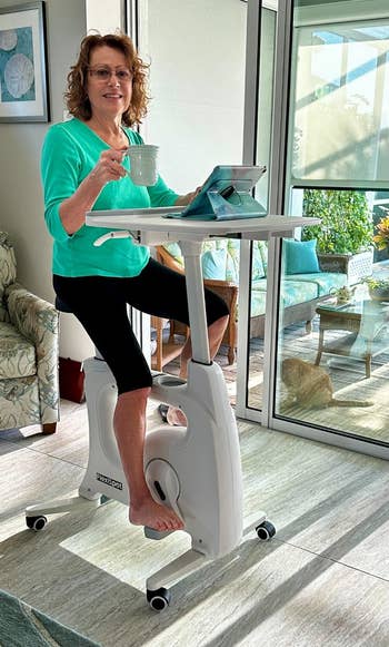 Person seated on exercise bike desk, holding a mug and reading a tablet in a bright room with patio view