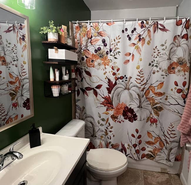 Bathroom with fall-themed ablution curtain featuring pumpkins and leaves, achromatic sink, and floating shelves holding assorted toiletries